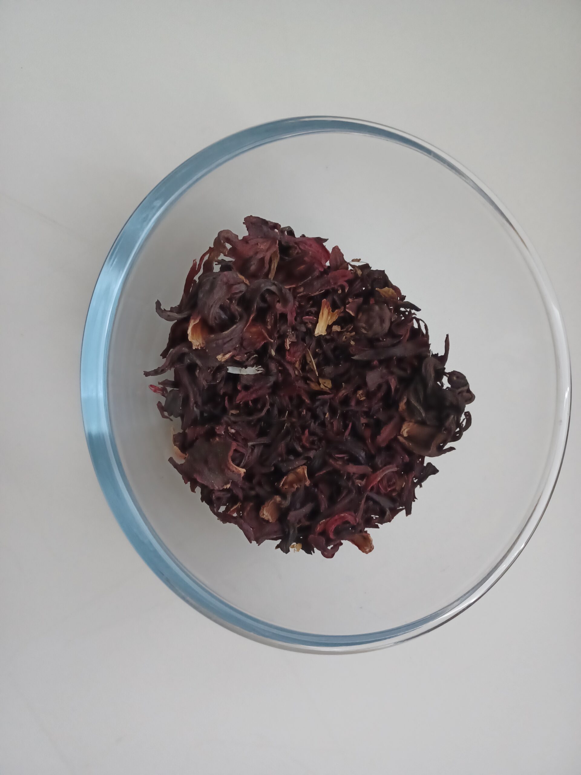 Hibiscus sabdariffa dried red calyces in transparent bowl on kitchen table showing concentrated anthocyanin pigment content in dried plant material where delphinidin and cyanidin secondary metabolites accumulate through phenylpropanoid biosynthesis as seed dispersal attraction chemistry