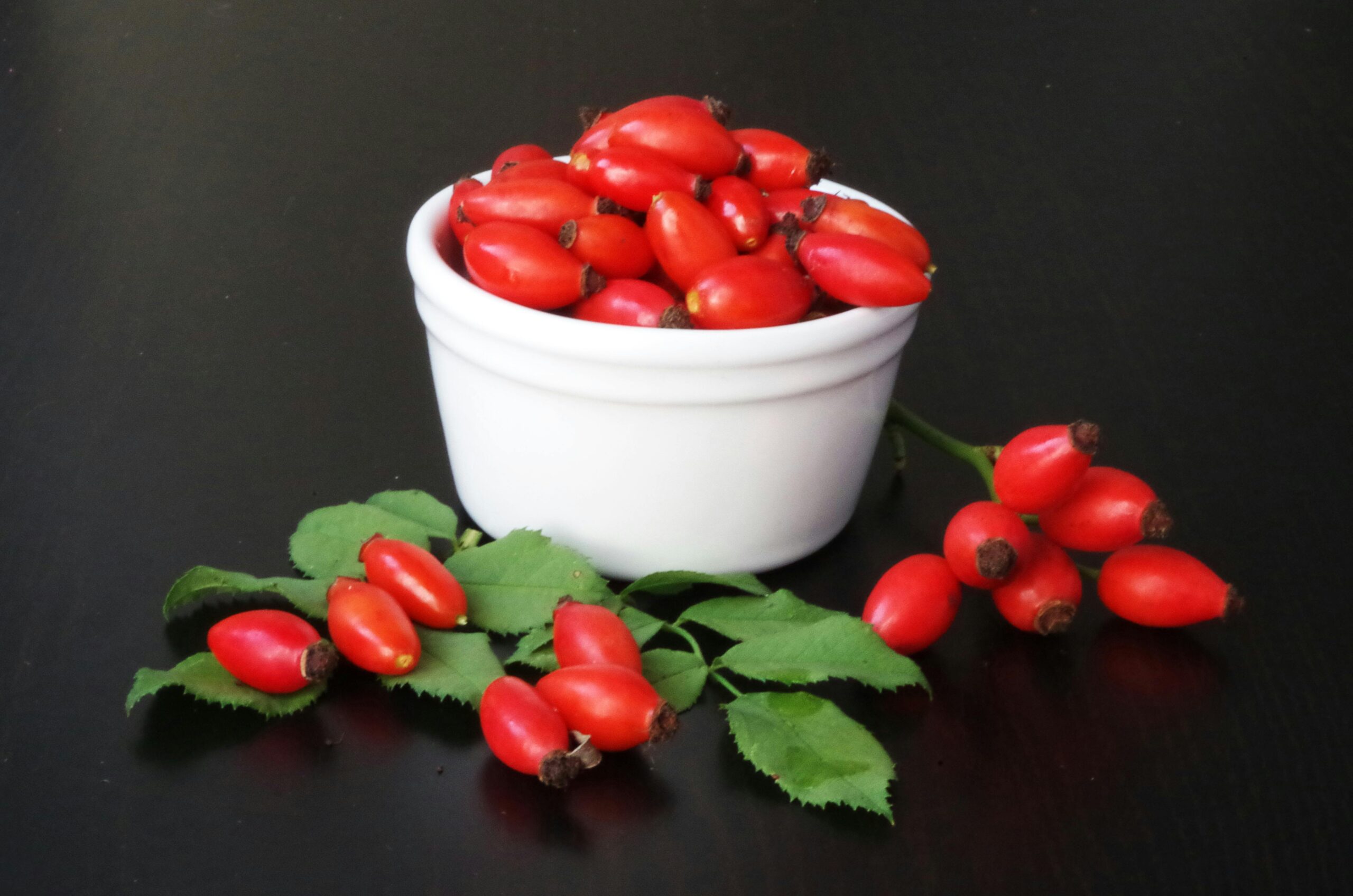 Rosa canina dog rose fresh red hip fruits in white bowl with leaves and stems showing accessory fruit structure where GOPO galactolipid vitamin C ascorbic acid carotenoid and polyphenol secondary metabolite compounds accumulate in flesh and seed tissue with documented anti-inflammatory and joint support activity