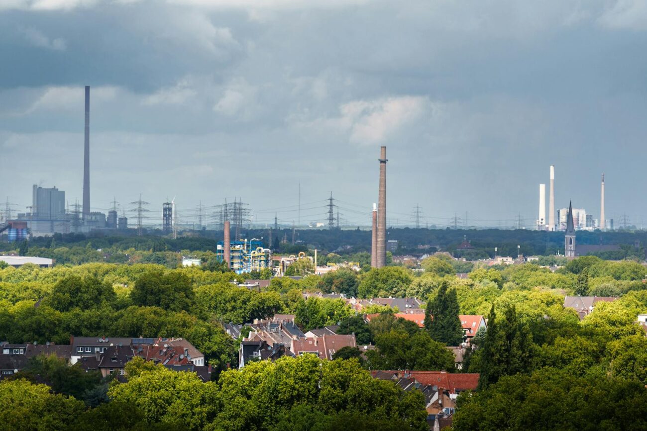 Urban tree canopy in residential area with industrial chimneys and factory emissions visible in background showing plants actively dealing with air pollution through antioxidant enzyme systems phenolic compound production and physical leaf surface barriers
