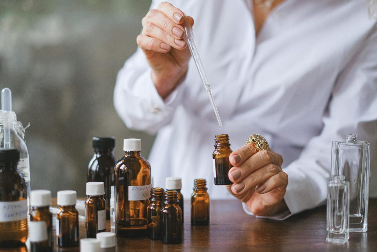Hands using glass dropper to blend essential oils from multiple amber bottles showing the precise measurement and compound combination process for DIY aromatherapy blends