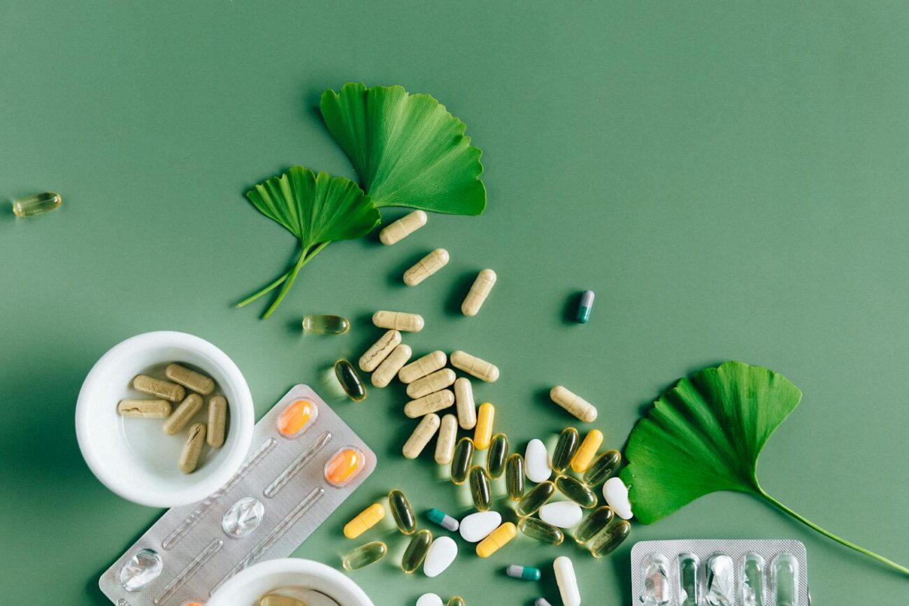 Herbal supplement capsules alongside pharmaceutical blister pack tablets with Ginkgo biloba leaves showing contrast between whole plant secondary metabolite preparations and isolated standardised pharmaceutical drug compounds both originating from plant chemistry