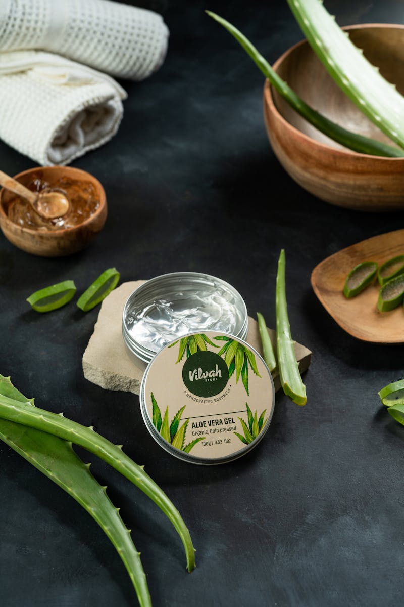 An open tin of organic aloe vera gel beside fresh aloe barbadensis leaves and cut pieces showing the plant source and prepared topical preparation
