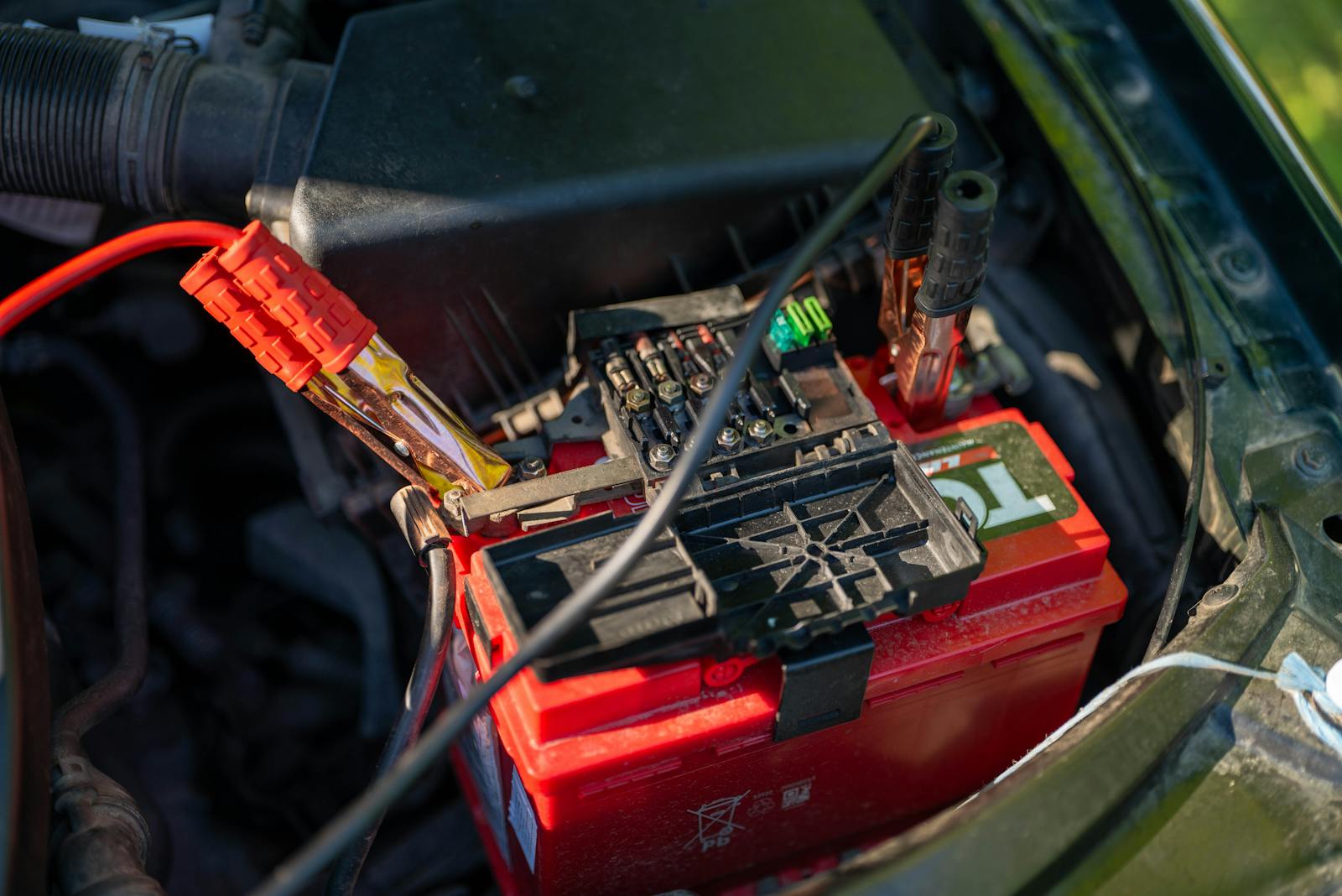 A red lead acid car battery in an engine bay with jump lead clamps connected to the terminals