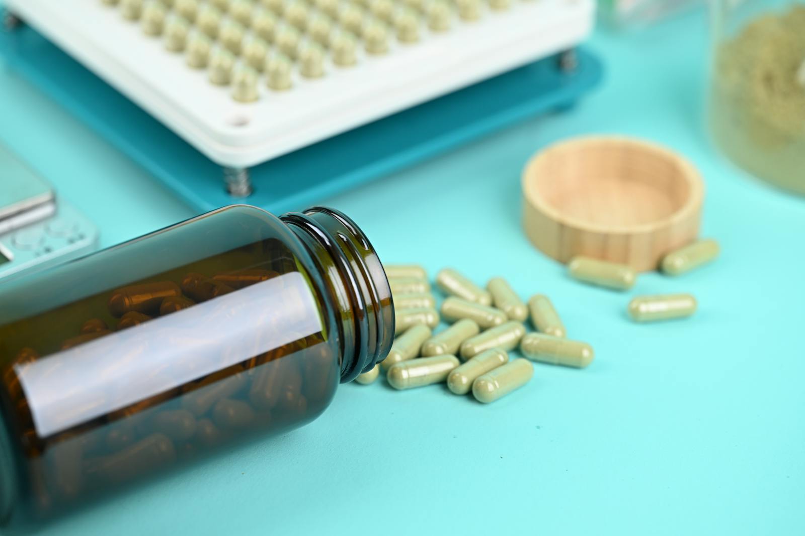 Green herbal supplement capsules spilling from an amber glass bottle with a capsule filling tray in the background showing supplement production quality context