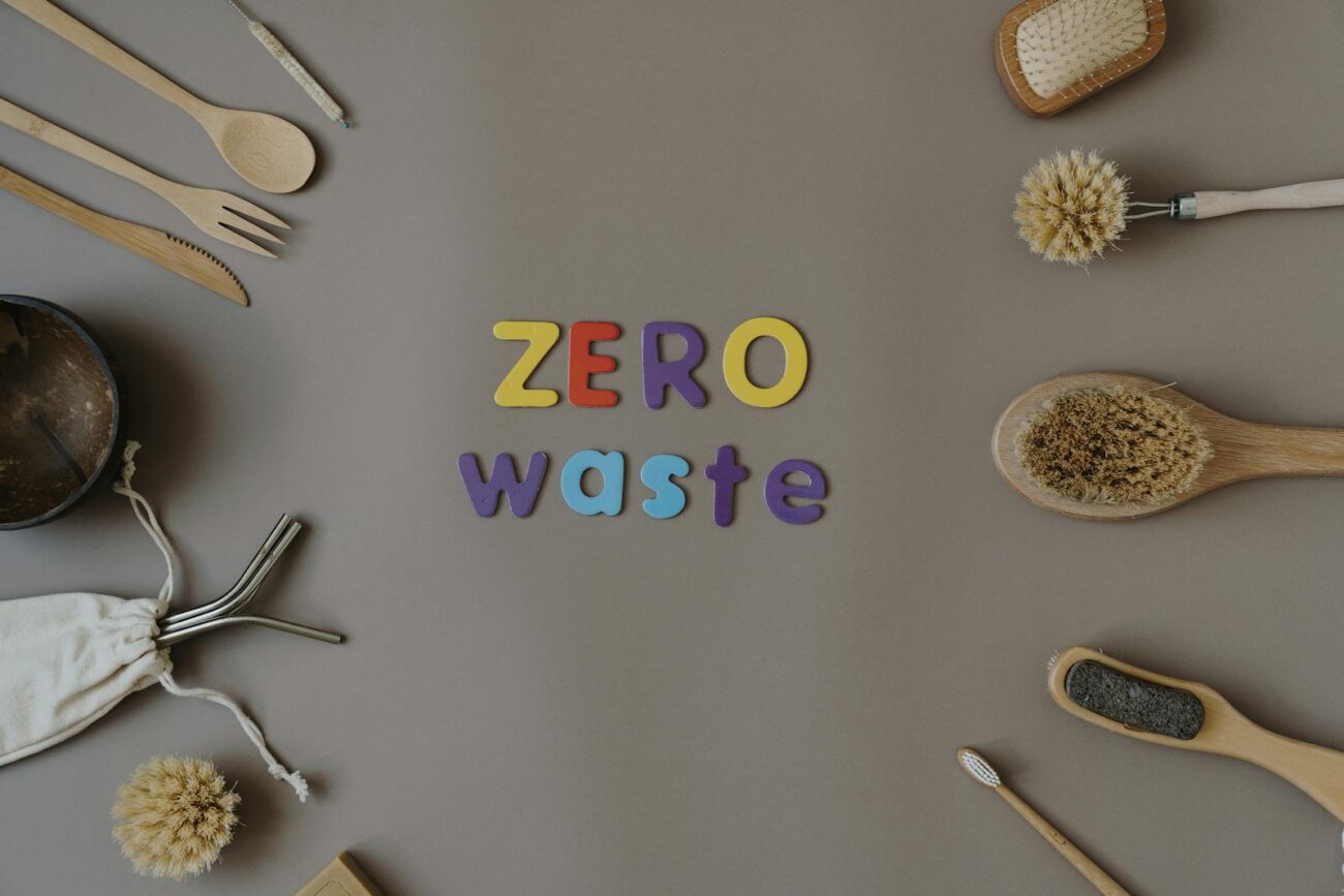 A flat lay of zero waste natural material alternatives including bamboo utensils natural brushes metal straws and a bamboo toothbrush arranged around the words zero waste