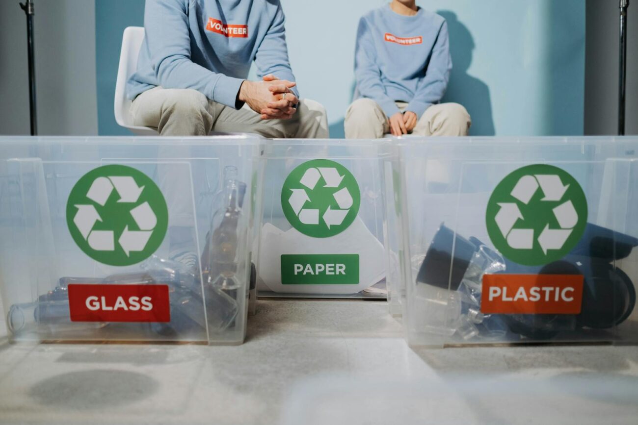 Two volunteers sitting behind labelled recycling bins for glass paper and plastic showing material stream separation in a circular economy system
