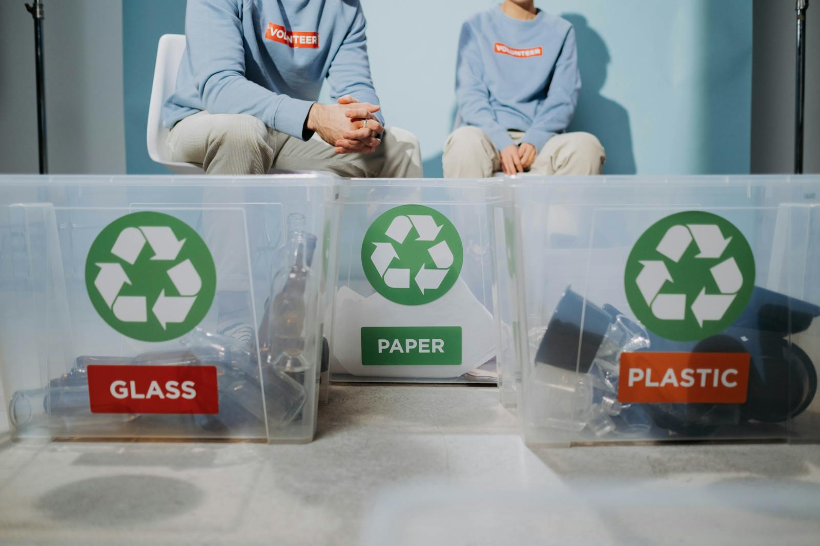 Two volunteers sitting behind labelled recycling bins for glass paper and plastic showing material stream separation in a circular economy system