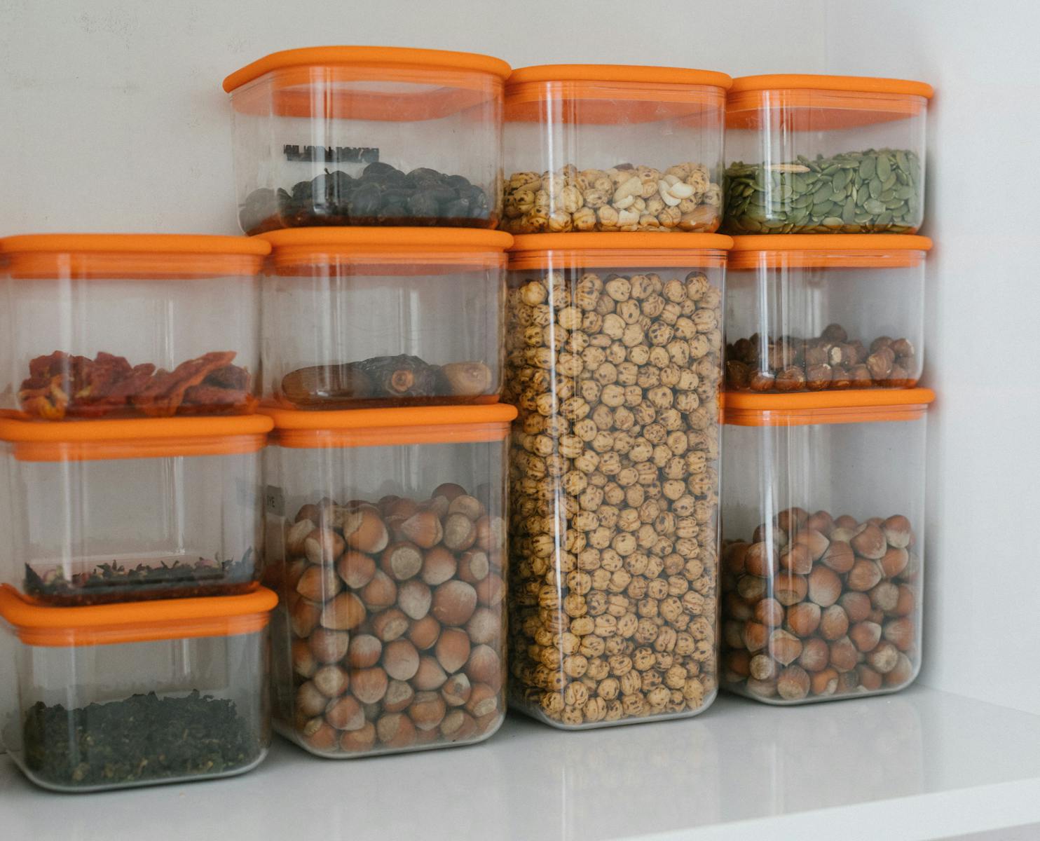 Clear plastic food storage containers with orange lids filled with nuts seeds and dried food on a kitchen shelf