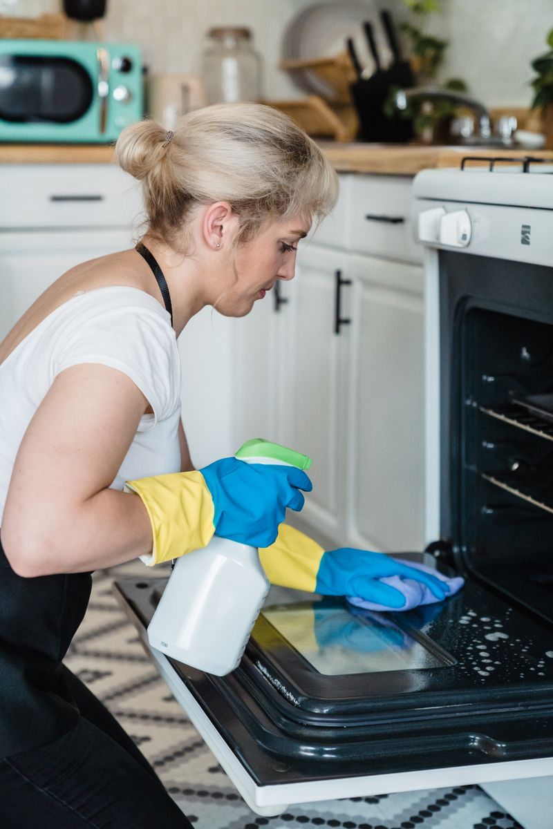A woman wearing rubber gloves cleaning an oven door with a spray bottle and cloth in a kitchen showing indoor cleaning where VOCs from conventional products accumulate