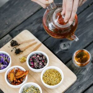 Hands brewing herbal tea in a glass teapot beside bowls of dried herbs and flowers
