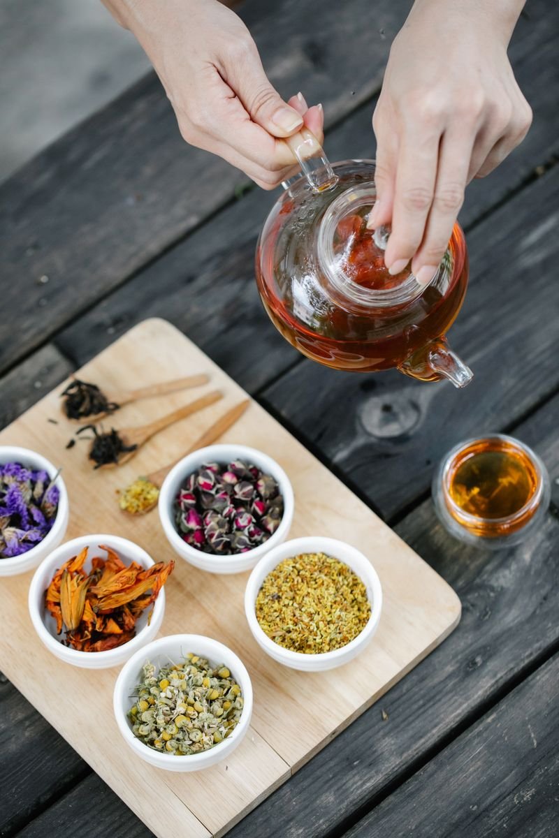 Hands brewing herbal tea in a glass teapot beside bowls of dried herbs and flowers